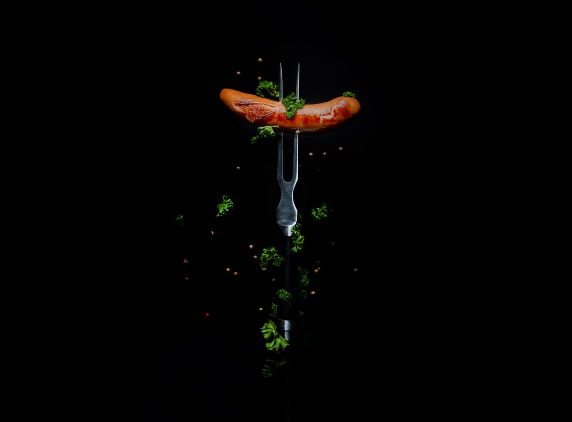 Eine Bratwurst auf einer Gabel gespießt vor schwarzem Glas